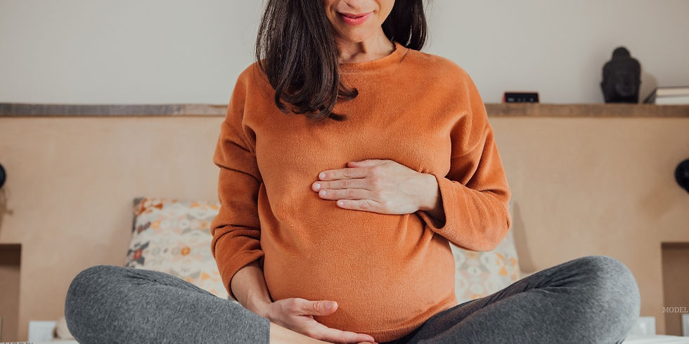 Pregnant woman sitting comfortably on a bed, resting her hands on her belly (model)